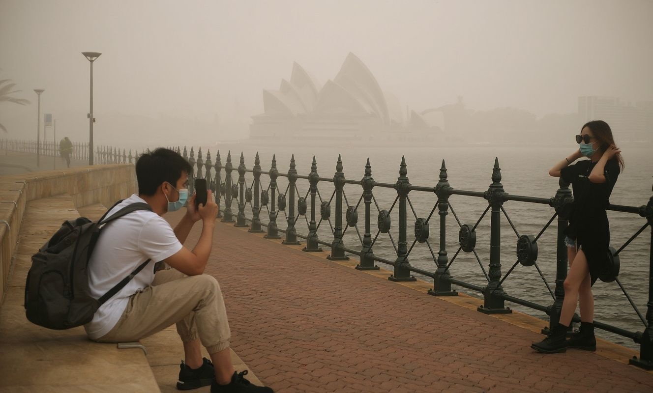 Tourists wearing masks take photos as the Opera House (back, C) is enveloped in haze caused by nearby bushfires, in Sydney on December 10, 2019. – Toxic haze blanketed Sydney on December 10 triggering a chorus of smoke alarms to ring across the city, as Australians braced for “severe” weather conditions expected to fuel deadly bush blazes. (Photo by PETER PARKS / )